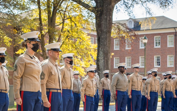 A formation of United States Marines in dress uniform (Service Charlie/Bravo) stands at attention during what appears to be a formal ceremony or parade. The Marines are wearing tan shirts, blue trousers with red blood stripes, and white covers (caps), with many wearing black face masks. The setting appears to be a military installation with a brick building and autumn trees visible in the background.