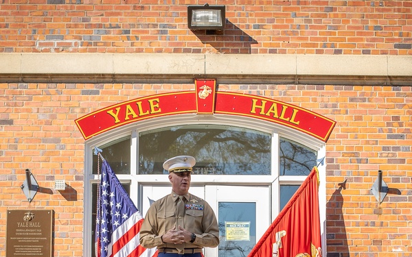 A U.S. Marine Corps officer in service uniform stands at attention in front of Yale Hall, a red brick building with a distinctive arched red-and-gold sign. He is flanked by the American flag on his left and a red U.S. Marine Corps unit flag on his right. The setting appears to be a formal military ceremony or official event at a Marine Corps installation.