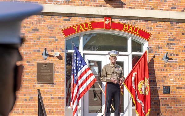 A senior U.S. Marine Corps officer in service uniform stands at the entrance of Yale Hall, a brick building on what appears to be a military installation, addressing personnel. The officer is flanked by the U.S. national flag and a red Marine Corps unit flag. A Marine in the foreground, wearing a white dress cover, is seen from behind observing the address.