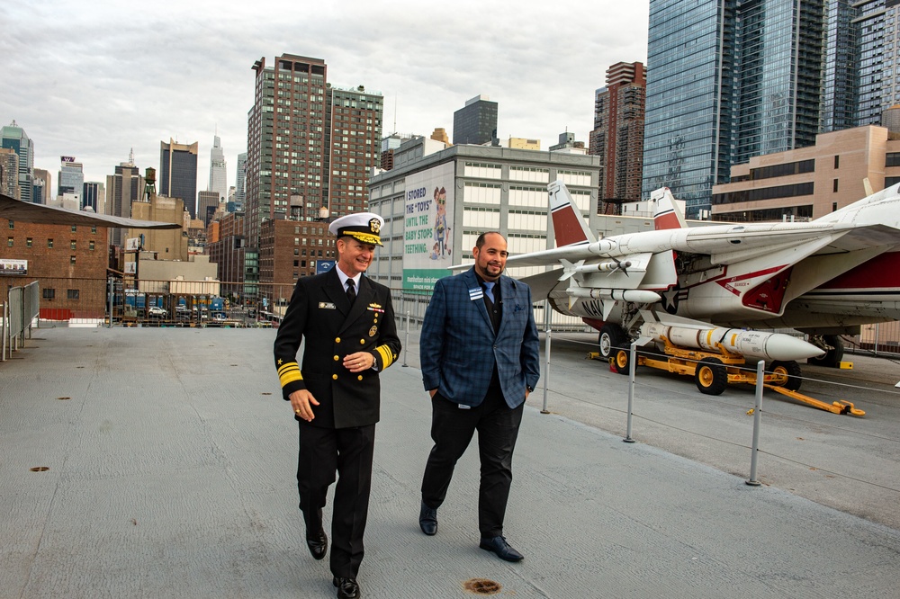 Two men walk together on the flight deck of what appears to be the USS Intrepid Sea, Air &amp; Space Museum in New York City. One man is dressed in a U.S. Navy dress uniform with gold rank insignia and a white officer's cap, while the other wears a blue plaid blazer. Military aircraft with missiles are visible on the deck, with the Manhattan skyline in the background.