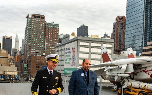 Two men walk together on the flight deck of what appears to be the USS Intrepid Sea, Air &amp; Space Museum in New York City. One man is dressed in a U.S. Navy dress uniform with gold rank insignia and a white officer's cap, while the other wears a blue plaid blazer. Military aircraft with missiles are visible on the deck, with the Manhattan skyline in the background.