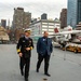Two men walk together on the flight deck of what appears to be the USS Intrepid Sea, Air &amp; Space Museum in New York City. One man is dressed in a U.S. Navy dress uniform with gold rank insignia and a white officer's cap, while the other wears a blue plaid blazer. Military aircraft with missiles are visible on the deck, with the Manhattan skyline in the background.