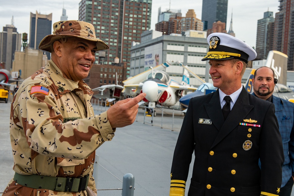 Two military officers are engaged in conversation on what appears to be the flight deck of the Intrepid Sea, Air &amp; Space Museum in New York City. One officer wears a desert camouflage uniform with a boonie hat and a U.S. flag patch, while the other wears a U.S. Navy dress uniform with gold insignia and a white officer's cap. Several vintage aircraft and the Manhattan skyline are visible in the background.