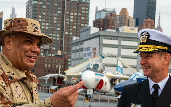 Two military officers are engaged in conversation on what appears to be the flight deck of the Intrepid Sea, Air &amp; Space Museum in New York City. One officer wears a desert camouflage uniform with a boonie hat and a U.S. flag patch, while the other wears a U.S. Navy dress uniform with gold insignia and a white officer's cap. Several vintage aircraft and the Manhattan skyline are visible in the background.