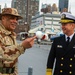 Two military officers are engaged in conversation on what appears to be the flight deck of the Intrepid Sea, Air &amp; Space Museum in New York City. One officer wears a desert camouflage uniform with a boonie hat and a U.S. flag patch, while the other wears a U.S. Navy dress uniform with gold insignia and a white officer's cap. Several vintage aircraft and the Manhattan skyline are visible in the background.