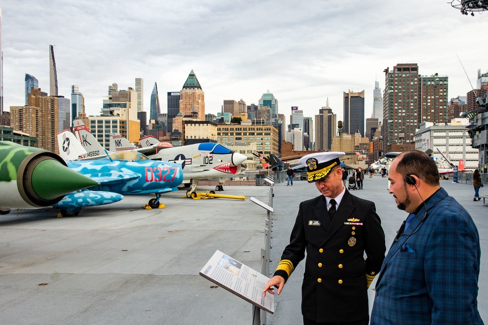 A U.S. Navy admiral in full dress uniform points to an informational placard while speaking with a civilian on the flight deck of what appears to be the Intrepid Sea, Air &amp; Space Museum in New York City. Several military aircraft are displayed on the deck, including a colorfully painted jet with 'D327' markings and another marked '102'. The Manhattan skyline is clearly visible in the background.