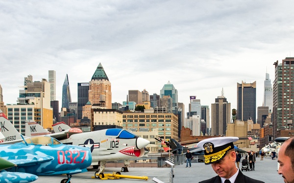 A U.S. Navy admiral in full dress uniform points to an informational placard while speaking with a civilian on the flight deck of what appears to be the Intrepid Sea, Air &amp; Space Museum in New York City. Several military aircraft are displayed on the deck, including a colorfully painted jet with 'D327' markings and another marked '102'. The Manhattan skyline is clearly visible in the background.