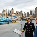 A U.S. Navy admiral in full dress uniform points to an informational placard while speaking with a civilian on the flight deck of what appears to be the Intrepid Sea, Air &amp; Space Museum in New York City. Several military aircraft are displayed on the deck, including a colorfully painted jet with 'D327' markings and another marked '102'. The Manhattan skyline is clearly visible in the background.