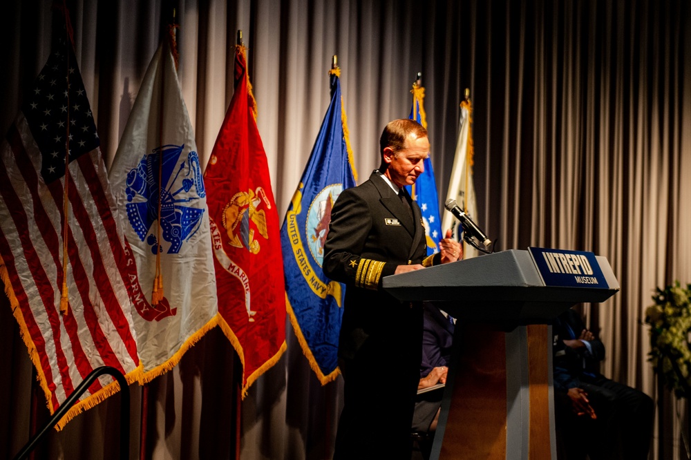 A senior U.S. Navy officer, identifiable by gold sleeve stripes indicating flag officer rank, speaks at a podium labeled 'Intrepid Museum.' The setting appears to be a formal military or commemorative ceremony, with multiple U.S. military service flags displayed behind the speaker. The flags visible include those of the U.S. Army, U.S. Marine Corps, U.S. Navy, U.S. Air Force, and the U.S. national flag.