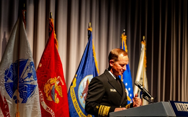 A senior U.S. Navy officer, identifiable by gold sleeve stripes indicating flag officer rank, speaks at a podium labeled 'Intrepid Museum.' The setting appears to be a formal military or commemorative ceremony, with multiple U.S. military service flags displayed behind the speaker. The flags visible include those of the U.S. Army, U.S. Marine Corps, U.S. Navy, U.S. Air Force, and the U.S. national flag.