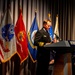 A senior U.S. Navy officer, identifiable by gold sleeve stripes indicating flag officer rank, speaks at a podium labeled 'Intrepid Museum.' The setting appears to be a formal military or commemorative ceremony, with multiple U.S. military service flags displayed behind the speaker. The flags visible include those of the U.S. Army, U.S. Marine Corps, U.S. Navy, U.S. Air Force, and the U.S. national flag.
