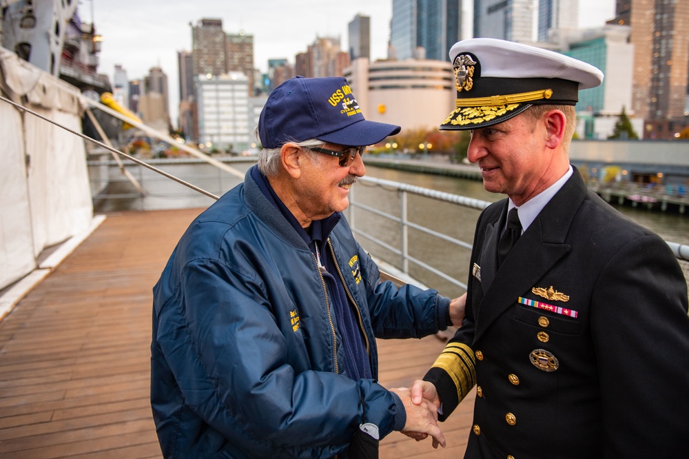 Two individuals are shaking hands on what appears to be the deck of a naval vessel or museum ship, with a New York City skyline visible in the background. One person is an older civilian wearing a blue USS-branded jacket and cap, while the other is a senior U.S. Navy officer in formal dress uniform with gold sleeve stripes and service ribbons. The setting suggests a ceremonial or commemorative event, likely near the Intrepid Sea, Air &amp; Space Museum on the Hudson River.