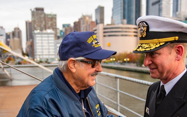 Two individuals are shaking hands on what appears to be the deck of a naval vessel or museum ship, with a New York City skyline visible in the background. One person is an older civilian wearing a blue USS-branded jacket and cap, while the other is a senior U.S. Navy officer in formal dress uniform with gold sleeve stripes and service ribbons. The setting suggests a ceremonial or commemorative event, likely near the Intrepid Sea, Air &amp; Space Museum on the Hudson River.