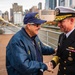 Two individuals are shaking hands on what appears to be the deck of a naval vessel or museum ship, with a New York City skyline visible in the background. One person is an older civilian wearing a blue USS-branded jacket and cap, while the other is a senior U.S. Navy officer in formal dress uniform with gold sleeve stripes and service ribbons. The setting suggests a ceremonial or commemorative event, likely near the Intrepid Sea, Air &amp; Space Museum on the Hudson River.