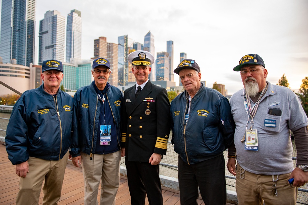 Five men pose for a group photo on what appears to be the deck of the USS Intrepid Sea, Air &amp; Space Museum in New York City. Four of the men are wearing navy blue 'USS Intrepid' caps and jackets, identifying them as veterans of the ship, while the central figure is dressed in a formal U.S. Navy officer's dress uniform. A modern Manhattan skyline is visible in the background.