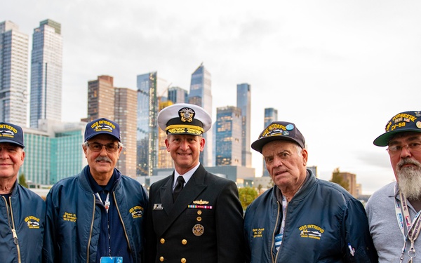 Five men pose for a group photo on what appears to be the deck of the USS Intrepid Sea, Air &amp; Space Museum in New York City. Four of the men are wearing navy blue 'USS Intrepid' caps and jackets, identifying them as veterans of the ship, while the central figure is dressed in a formal U.S. Navy officer's dress uniform. A modern Manhattan skyline is visible in the background.