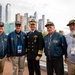 Five men pose for a group photo on what appears to be the deck of the USS Intrepid Sea, Air &amp; Space Museum in New York City. Four of the men are wearing navy blue 'USS Intrepid' caps and jackets, identifying them as veterans of the ship, while the central figure is dressed in a formal U.S. Navy officer's dress uniform. A modern Manhattan skyline is visible in the background.