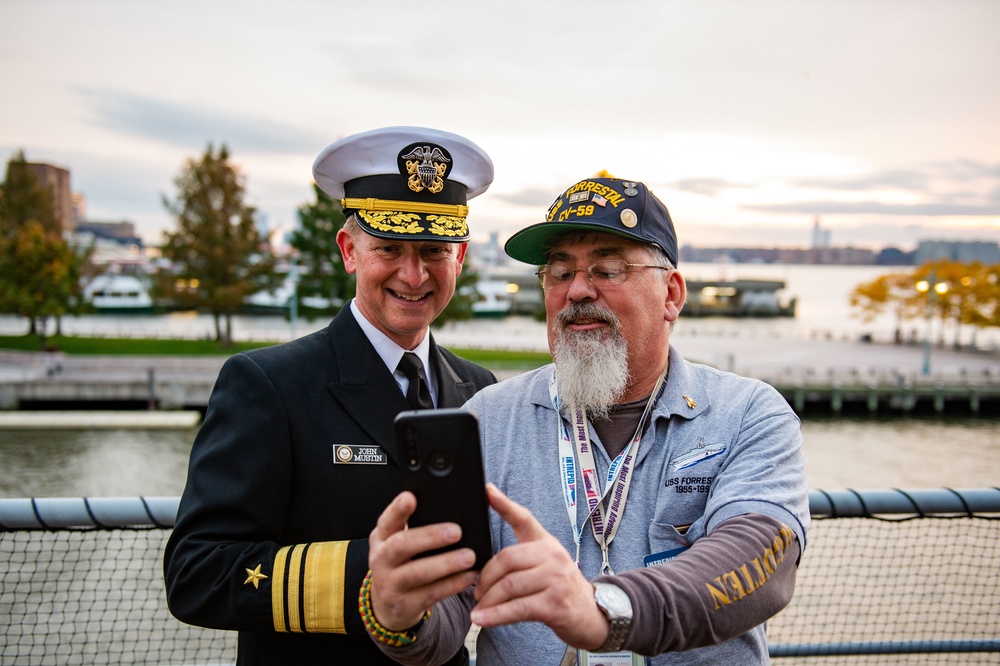 A U.S. Navy officer in dress uniform, identified by his name tag as John Mustin, poses for a selfie with a civilian man wearing a USS Forrestal (CV-59) veteran's cap and shirt. The two men are looking at a smartphone held by the civilian, both appearing engaged and smiling. The setting appears to be an outdoor waterfront location, likely during a naval or veterans' event.
