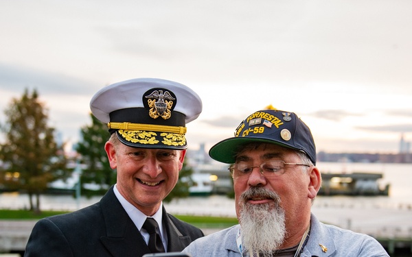 A U.S. Navy officer in dress uniform, identified by his name tag as John Mustin, poses for a selfie with a civilian man wearing a USS Forrestal (CV-59) veteran's cap and shirt. The two men are looking at a smartphone held by the civilian, both appearing engaged and smiling. The setting appears to be an outdoor waterfront location, likely during a naval or veterans' event.