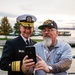 A U.S. Navy officer in dress uniform, identified by his name tag as John Mustin, poses for a selfie with a civilian man wearing a USS Forrestal (CV-59) veteran's cap and shirt. The two men are looking at a smartphone held by the civilian, both appearing engaged and smiling. The setting appears to be an outdoor waterfront location, likely during a naval or veterans' event.