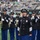 New York Jets Salute to Service Game 11/14/21 A U.S. Army military band is performing on a football field, likely during a pre-game or halftime ceremony at an NFL stadium. A female soldier in the foreground, wearing a senior NCO rank insignia and service medals, is singing into a microphone while band members behind her play saxophones, brass instruments, and a sousaphone. The stadium in the background shows spectators in the stands, with green and blue team colors visible.