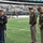 New York Jets Salute to Service Game 11/14/21 Three U.S. Army officers in dress uniforms are participating in a military ceremony on the field of MetLife Stadium, home of the New York Jets. One officer on the left, wearing the Army Service Uniform (blue), appears to be reading from a binder, while two officers in Army Green Service Uniforms stand with heads bowed in a reverent posture. The event appears to be a 'Salute to Service' military appreciation ceremony during an NFL game.