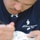 Navy Wounded Warriors Take Part in Ceramics in Honor of Warrior Care Month A person wearing a navy blue Under Armour shirt with an embroidered 'Navy Wounded Warrior NRH' logo is leaning forward and writing or drawing on a white object. The shirt features a lighthouse graphic above the text. The subject's face is partially visible but out of focus, with the shirt logo being the sharpest element in the frame.