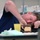 Navy Wounded Warriors Take Part in Ceramics in Honor of Warrior Care Month A man in a navy blue shirt is lying with his head resting on a table, closely examining a small clay or ceramic sculpture. He appears to be working on or inspecting a handmade mug or cup-shaped object with a light-colored rectangular panel on it. The setting appears to be a classroom or workshop environment.
