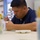 Navy Wounded Warriors Take Part in Ceramics in Honor of Warrior Care Month A man wearing a navy blue Under Armour shirt is focused on painting or writing on a white ceramic mug at a table. He is holding a fine-tipped brush or pen and appears to be carefully applying lettering or a design to the mug. The setting appears to be a paint-your-own-pottery or ceramic arts studio.