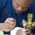 Navy Wounded Warriors Take Part in Ceramics in Honor of Warrior Care Month A man wearing a navy blue t-shirt with yellow anchor and chevron graphics is carefully painting or decorating a white ceramic mug using a fine-tipped brush. He is leaning in closely and concentrating intently on his work. A small green container, likely holding paint or water, is visible in the lower left corner.