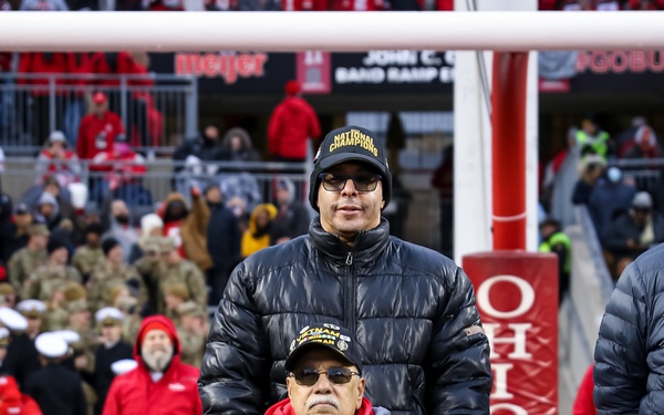 Two individuals are on the field at Ohio State University's football stadium during what appears to be a veteran recognition ceremony. One person is seated in a wheelchair wearing a Vietnam veteran cap and red Ohio State apparel, while another person stands behind pushing the wheelchair. Military personnel and a large crowd are visible in the background.