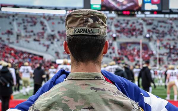 A soldier with the name tape 'KARMAN' on their Operational Camouflage Pattern (OCP) uniform stands on the sideline of a packed American football stadium, holding an unfolded American flag across their back. The soldier is flanked by other uniformed military personnel, suggesting a ceremonial flag presentation during a pre-game or halftime event. The stadium is filled with spectators predominantly wearing red, and players are visible on the field.