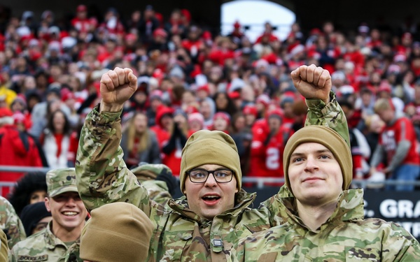 A group of military personnel in various uniforms are enthusiastically cheering at what appears to be a college football game. Two individuals in Army Combat Uniforms (ACU) with tan beanies are raising their fists in celebration, while others in Navy dress whites are visible in the foreground. The stadium behind them is packed with fans predominantly wearing red, suggesting a home team color scheme.