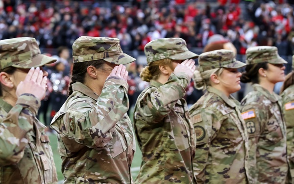 A group of U.S. Army soldiers, all appearing to be women, stand in formation on a football field and render a salute. They are wearing Operational Camouflage Pattern (OCP) uniforms with American flag patches visible on their right shoulders. The setting appears to be a military appreciation ceremony during a college or professional football game, with a packed stadium crowd visible in the background.