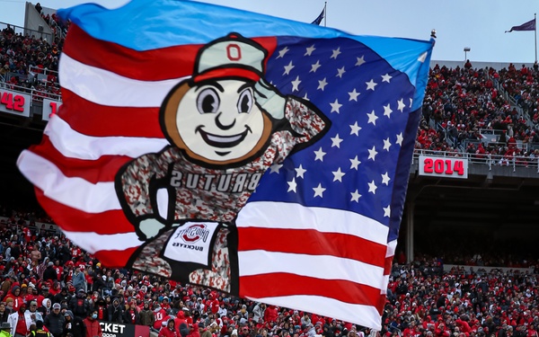 The Ohio State University mascot Brutus Buckeye is waving a large flag that combines the American flag design with the Brutus Buckeye character in military camouflage attire. The scene takes place at Ohio Stadium (The Horseshoe) during a football game, with a packed crowd of fans predominantly wearing red and gray. The stadium is filled to capacity with spectators visible in multiple seating tiers.