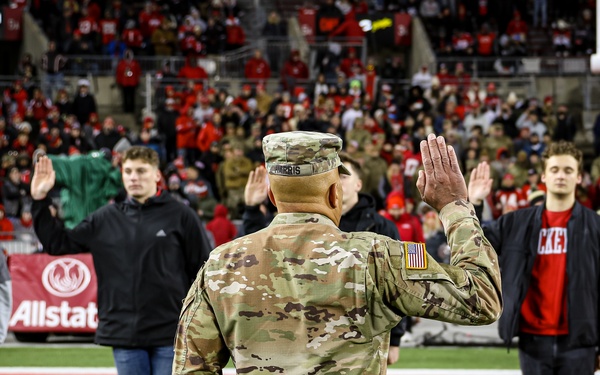A U.S. Army soldier in MultiCam/OCP camouflage uniform is conducting a military enlistment or oath of office ceremony on a football field, facing a group of civilians with raised right hands. The ceremony is taking place at what appears to be an Ohio State University football game at Ohio Stadium, with a large crowd of fans dressed predominantly in red visible in the background. An Allstate sponsorship banner is visible on the field perimeter.