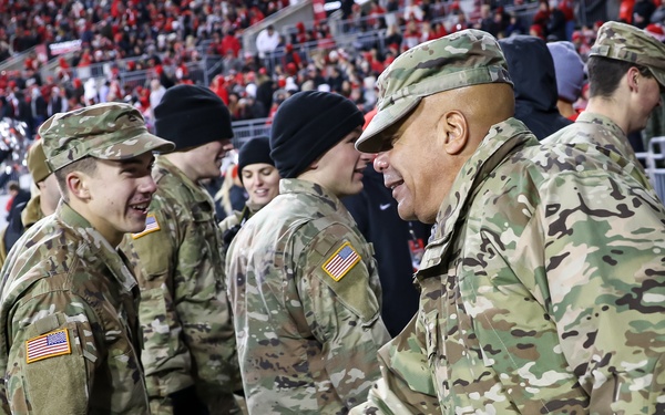 A group of U.S. Army personnel in MultiCam (OCP) camouflage uniforms are gathered on what appears to be a football field sideline during a night game, with a packed stadium crowd visible in the background. A senior soldier on the right is shaking hands with a younger soldier on the left, who is smiling. The setting suggests a military appreciation or recognition ceremony at a sporting event.