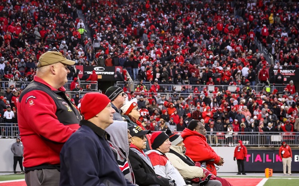 A group of individuals, several in wheelchairs, are lined up on the sideline of a football field during what appears to be a recognition ceremony. The stadium is packed with fans predominantly wearing red, suggesting a major college football game, likely Ohio State given the team colors and branding visible. The group appears to include veterans or honorees being recognized during a halftime or pre-game ceremony.