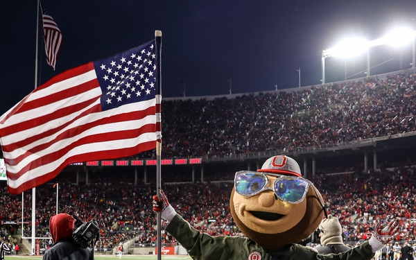 Brutus Buckeye, the official mascot of Ohio State University, is shown on the sideline of a night football game at Ohio Stadium (The Horseshoe) in Columbus, Ohio. The mascot is dressed in an olive green flight suit and large reflective aviator sunglasses, holding an American flag. The stadium is packed with fans predominantly wearing red, and the field's end zone is visible in the background.