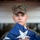A young U.S. Marine in MARPAT woodland digital camouflage uniform holds a folded American flag at chest level with a solemn expression. The image appears to be taken during a formal military ceremony, likely a flag presentation or memorial service. Additional Marines in matching uniforms are partially visible on either side.