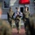 Three U.S. Marines in MARPAT digital camouflage uniforms are carrying a folded American flag during what appears to be a formal military ceremony. The center Marine holds the flag against their chest while flanked by two others, all marching in formation along a brick pathway. The setting appears to be an official military installation with a formal building entrance visible in the background.