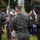 Three U.S. Marines in MARPAT digital camouflage uniforms are conducting a formal flag ceremony at a flagpole, handling a folded American flag. A large brass bell is visible at the top of the flagpole. Additional uniformed personnel and what appears to be a military band are visible in the background.
