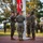 A formal military flag-raising ceremony is taking place on what appears to be a U.S. Marine Corps installation. Several Marines in MARPAT camouflage uniforms are actively raising a red flag at a flagpole while others render salutes. A military band with brass and woodwind instruments is assembled in the background providing ceremonial music.