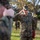 A U.S. Marine in MARPAT woodland camouflage uniform is rendering a salute during what appears to be a formal military ceremony outdoors. He is holding a red folder/binder in his left hand while saluting with his right. A military band is visible in the background, and another Marine in the foreground is also saluting.