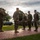 A military ceremony is taking place outdoors on a brick plaza, likely at a U.S. Marine Corps installation. A senior officer on the left is reading from a document to a formation of Marines standing at attention. The setting includes a flagpole with a bell, green lawn, and trees in the background.