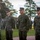 A group of U.S. Marines in MARPAT woodland digital camouflage uniforms are gathered outdoors on a grassy area, appearing to be in a formation or inspection setting. A senior Marine on the left appears to be addressing or inspecting several Marines standing at attention. The setting appears to be a military base with trees and a body of water visible in the background.