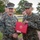 Two U.S. Marines in MARPAT woodland camouflage uniforms are shaking hands during what appears to be a formal award or recognition ceremony. The senior Marine, identified by his name tape as 'DONOVAN,' is presenting a red official folder bearing the U.S. Marine Corps emblem to a junior Marine. The ceremony is taking place outdoors on a military installation with other Marines visible in formation in the background.