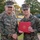 Two U.S. Marines in MARPAT digital camouflage uniforms are posing for a formal photograph outdoors. The senior Marine, identified by his name tape as 'DONOVAN,' is shaking hands with a younger Marine who is holding a red presentation folder bearing the U.S. Marine Corps emblem. This appears to be an award or commendation ceremony taking place on a military installation.