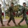 Three U.S. Marines in MARPAT woodland camouflage uniforms march in formation across a brick pathway during what appears to be a formal military ceremony. Each of the three lead Marines carries a red folder or binder. In the background, a military band with brass instruments is assembled on a grass field alongside additional uniformed personnel.