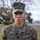 A U.S. Marine in woodland MARPAT digital camouflage uniform stands at attention outdoors. The name tape reads 'BURNS' and the branch tape reads 'U.S. MARINES,' with rank insignia visible on the collar. The subject appears young and is photographed in a formal portrait-style composition against a blurred outdoor background.
